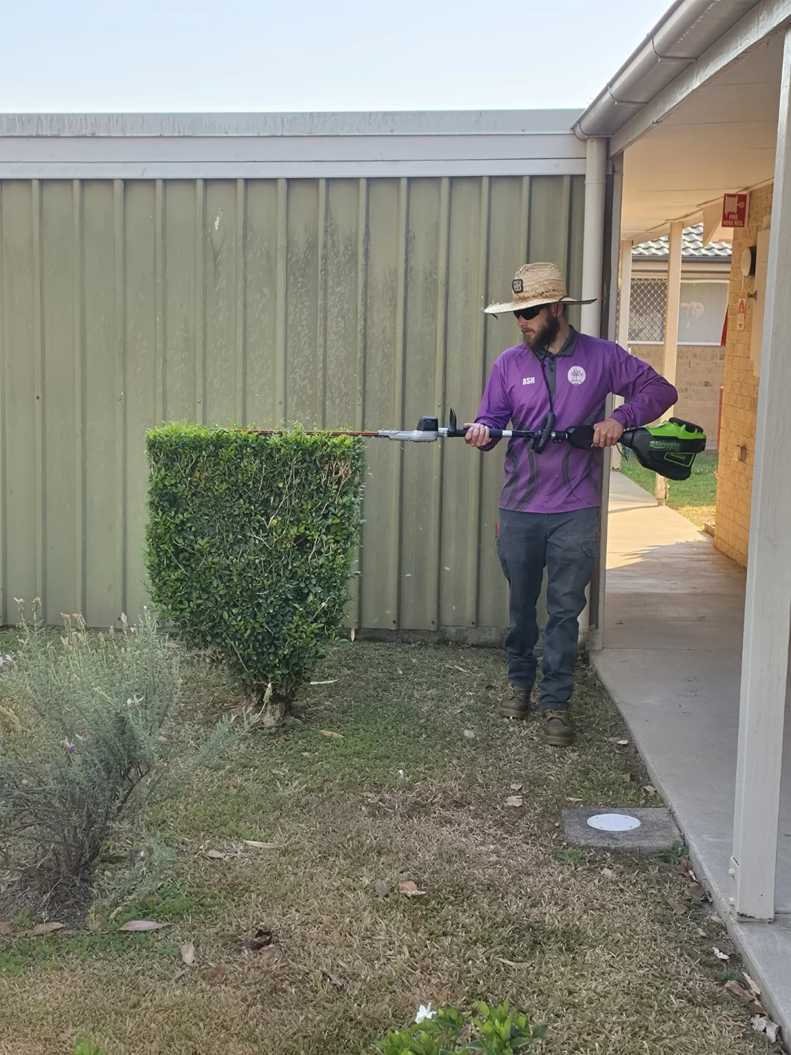 An Excel team member trimming the top of a rectangular hedge beside a shared walkway
