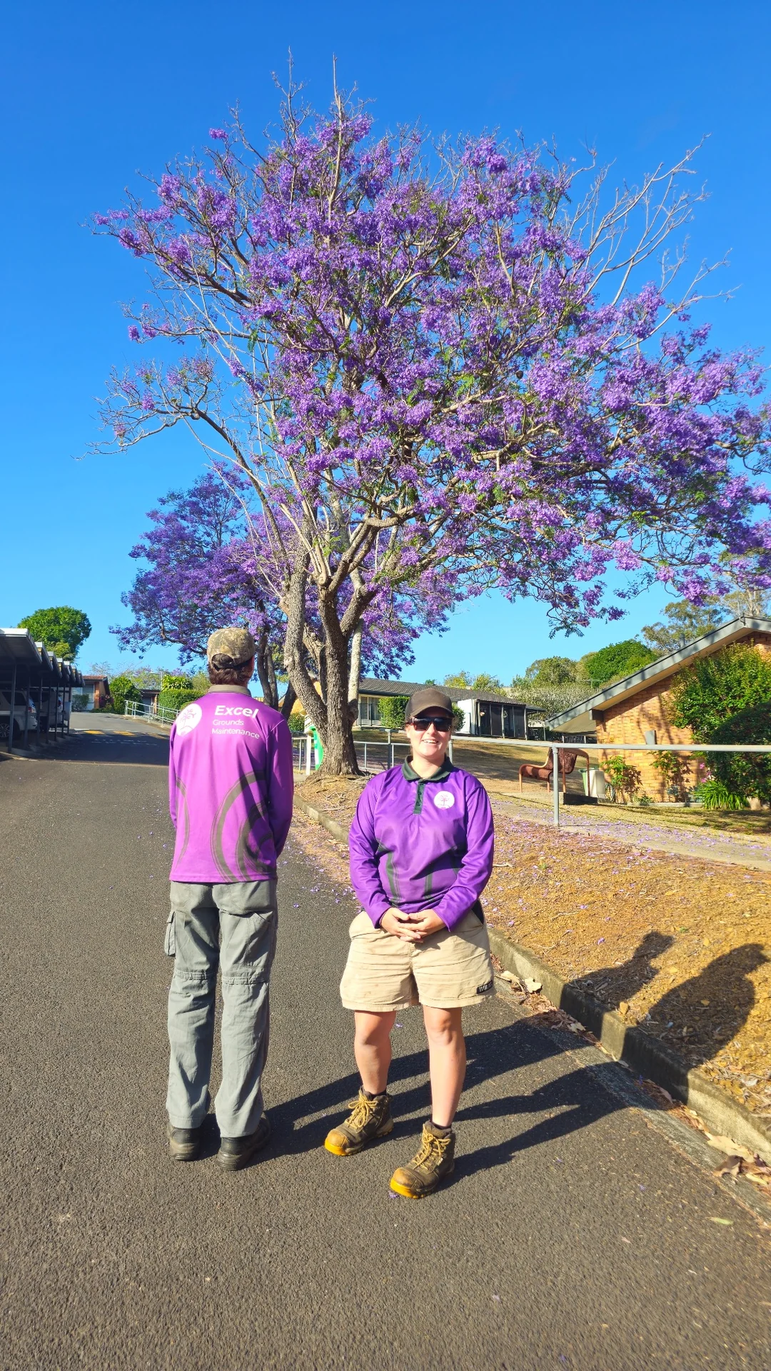 Two Excel team members beneath a flowering purple jacaranda in a village