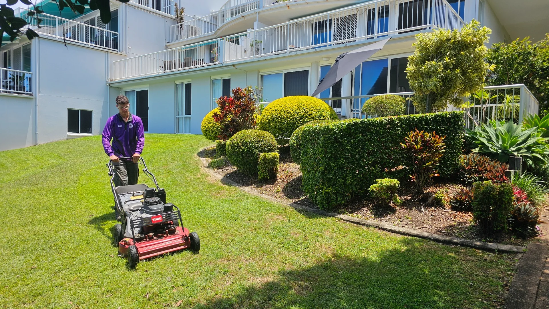 An Excel team member mowing beside shaped hedges and village apartments